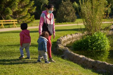 Maskeli bir anne çocuklarını yüzme havuzunda gezdirir. Parkta iki çocuklu bir kadın. Yeni normal. Grup oturumu