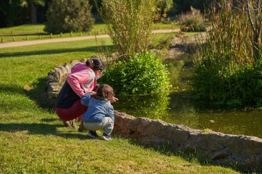 Maskeli anne havuza bakarken çömelme pozisyonunda yürür. Çocuklu bir kadın el ele tutuşuyor. Aile toplantısı. Grup oturumu