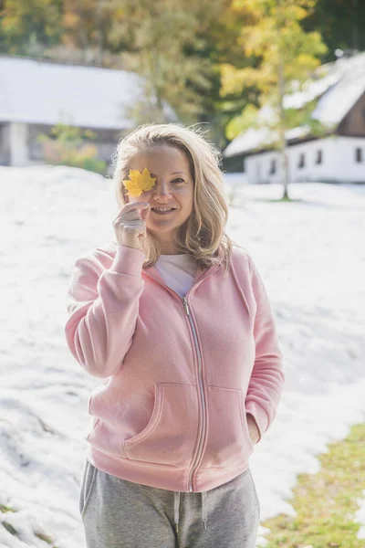 Attractive blonde hides her face behind an autumn leaf. Snow-covered alpine village in autumn.