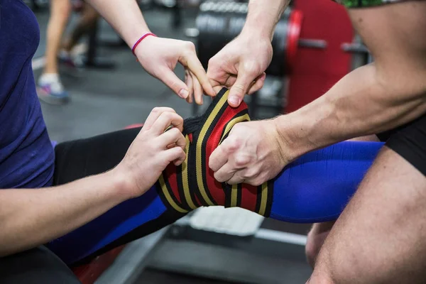 Sporcu, spor salonunda eşinin dizini elastik bandajla bandajlıyor. Bir halterle çömel..