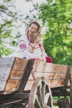 Nakışlı giysiler içindeki güzel köylü kadın kızıyla birlikte at arabasında oturuyor.