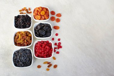 Farklı kurutulmuş meyveler: kuru erik, kuru üzüm, kuru kayısı ve kirazlar seramik bir kasede açık arkaplanda, üst manzara.