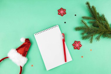 Noel kompozisyonu, istek listesi konsepti. Yeşil arka planda kalem, ladin dalları, süslemeler ve Noel Baba şapkaları olan defter. Düz yatış, üst görünüm.