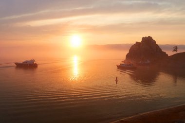 Baykal Gölü üzerinde renkli gün batımı. Şaman kayasının yakınındaki Khuzhir Körfezi manzarası. Körfezde turistli tekne ve yatlar dinleniyor. Sörf tahtasında küreği olan bir kadın var.