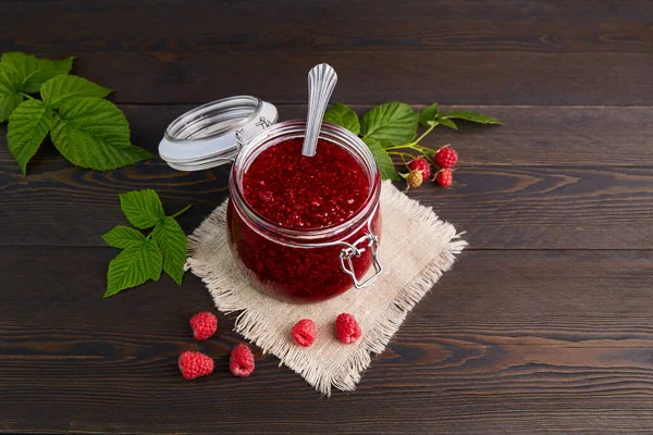 Raspberry jam in a glass jar on a linen napkin on a dark wooden background. Conservation and summer harvesting. — Stock Photo, Image