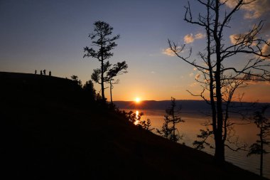İnsanlar bir kayanın üzerinde durup gün batımını seyrediyorlar. Turuncu bir günbatımında insan ve ağaç siluetleri.