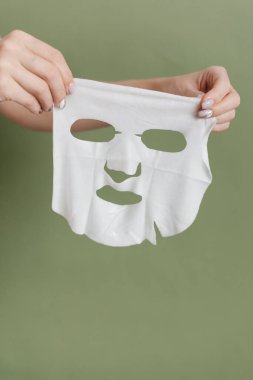 Bir kadının elleri beyaz kumaştan yapılmış kozmetik bir yüz maskesi tutuyor. Yeşil arka planda kozmetik yüz maskesi.