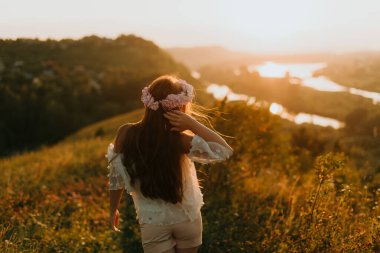 Batan güneşin altında güzel bir kız çimlerin üzerinde yürüyor. Çiçek çelengi