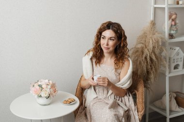 Bir kadın elinde bir fincan kahveyle dinleniyor. Masanın yanında güzel çiçekler ve kurabiyeler var.