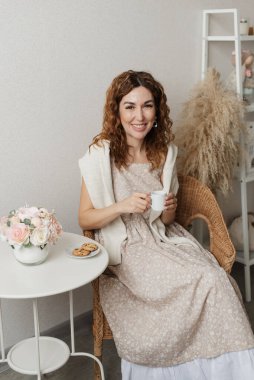 Kadın elinde beyaz bir fincan kahve tutuyor ve neşeyle kameraya bakıyor.