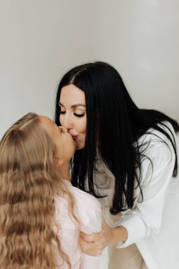 Güzel, genç, esmer bir anne beyaz arka planda uzun sarı saçlarıyla kızını öpüyor.