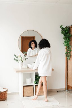 Beyaz kazaklı Afrika kıvırcık saçlı bir kadın yuvarlak bir aynada beyaz bir duvar arkasına bakıyor.