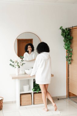 Beyaz kazaklı, kıvırcık saçlı, çıplak ayaklı, yuvarlak aynaya bakan bir kadın.