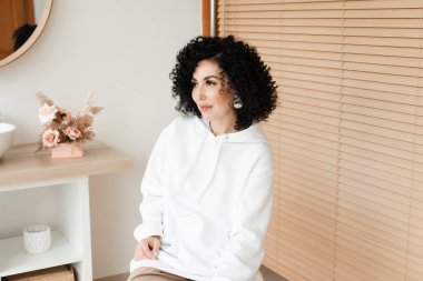 Beyaz kazaklı, kıvırcık saçlı esmer bir kadın gülümsüyor ve yan tarafa bakıyor.