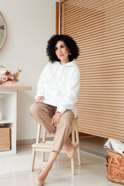 Beyaz kazaklı, bej pantolonlu, Afro kıvırcık saçlı bir kadın bir sandalyeye oturur ve yan tarafa bakar.