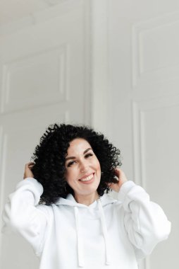 Beyaz kazaklı, siyah kıvırcık saçlı bir kadın beyaz bir duvara yaslanmış gülüyor ve saçlarına dokunuyor.