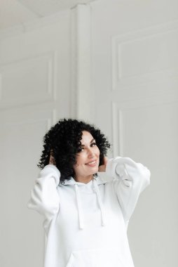 Beyaz kazaklı bir kadın gülümsüyor ve iki eliyle kıvırcık saçlarına dokunuyor.