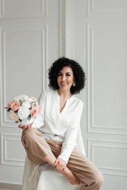 Çıplak ayakla gülümseyen kıvırcık kadın sandalyeye oturur ve elinde yuvarlak bir buket çiçek tutar.