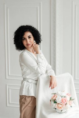 Afrika bukleleri olan bir kadın beyaz bir duvar arkasında bir buket çiçeğin yanında duruyor.