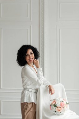 Afrikalı bukleli gülümseyen bir kadın beyaz bir duvar arkasında bir buket çiçeğin yanında duruyor.