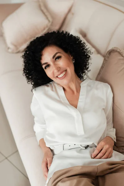 Curly woman smiling with teeth and lying on a pink sofa in a white blouse