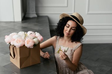 Elbise ve hasır şapkalı bir kadın elinde süs çiçekleri tutuyordu.