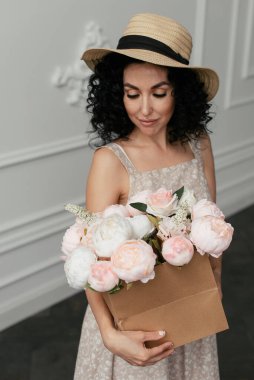 Pembe ve beyaz şakayıklar bir el sanatları kutusunda, saman şapkalı kıvırcık bir esmerin ellerinde.