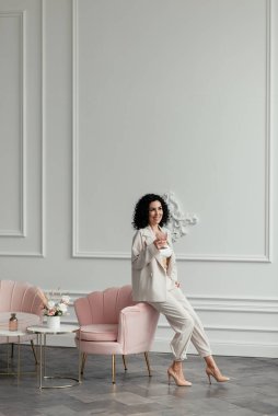 Koyu kıvırcık saçlı bir kadın pembe bir koltuğa oturmuş pembe bir bardak tutuyor.