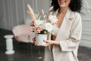 Takım elbiseli kıvırcık saçlı bir kadın elinde güzel bir buket çiçekle gülüyor.