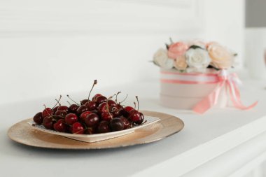 Beyaz bir masada bir tabak kiraz, arka planda pembe bir kutuda çiçekler.