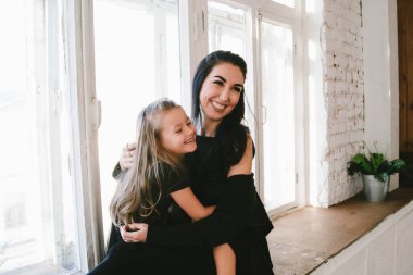 Gülümseyen siyah elbiseli anne ve kız pencerenin yanında sarılıyorlar.