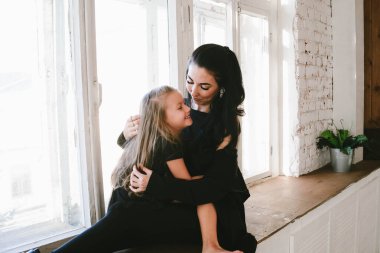 Siyah elbiseli şık esmer anne kızını pencerenin yanında kucaklıyor.