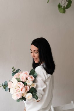 Beyaz gömlekli genç esmer bir kız duvarın yanında elinde büyük bir buket çiçek tutuyor.