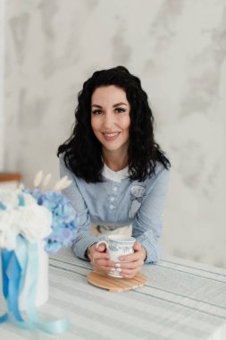 Siyah dalgalı saçlı, mavi gömlekli, elinde kupa tutan güzel esmer kız.