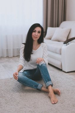 Beyaz kazaklı ve kot pantolonlu güzel esmer kız evde yerde oturuyor.