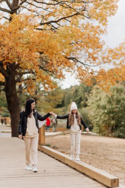 Şık bir anne ve kızı sonbahar parkında el ele yürür.
