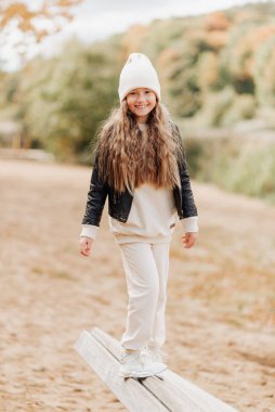 Beyaz şapkalı ve siyah deri ceketli şirin küçük kız sonbahar parkında oynaşıyor.