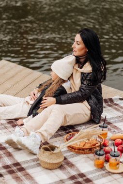 Siyah deri ceketli şık esmer anne, sonbahar pikniğinde kızına sarılıyor.