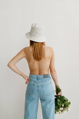 Sırtında kot pantolon ve beyaz arka planda panama şapkası olan çıplak bir kız.