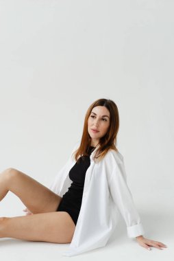 Beyaz tişörtlü, çıplak bacaklı güzel bir kız sade bir arka planda yerde oturuyor.