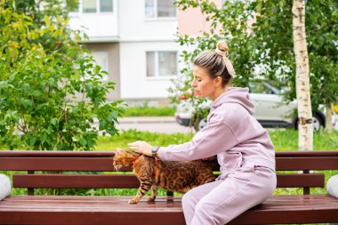 Dışarıda Bengal kedisiyle gezen bir kadın. Hayvan bakımı kavramı, koşumlu kedi, günlük yürüyüş, koşu, antrenman ve sevimli evcil hayvanlarla yapılan tüm açık hava eğlenceleri. Kız evcil hayvan sahibi kedisini taşıyor, oynuyor, kedisine aşık.