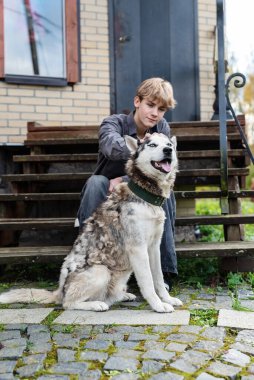 Genç beyaz erkek, tuğla evin önünde iri yarı bir köpekle basamaklarda oturuyor. Çocuk köpeğiyle bakım, sevgi ve ilgi, duygusal bağlantı, güven ve arkadaşlık yoluyla bağ kuruyor..