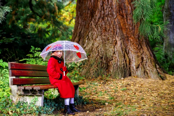 Menina bonito com guarda-chuva sentado sob o grande pinetree — Fotografia de Stock