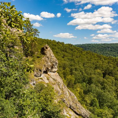 Yeşil ormanlı manzara ve mavi gökyüzünün altında bulutlu yüksek kayalık dağ