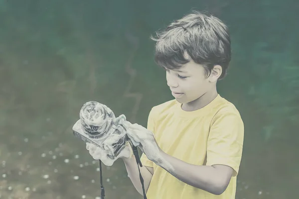 一个穿着黄色T恤和短裤的男孩站在大自然的水边 用肥皂和泡沫洗相机 概念小孩是个欺负人的人 电影效果 图库图片