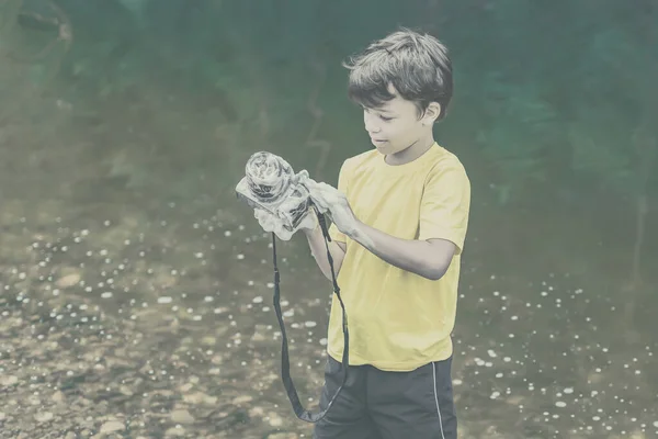 一个穿着黄色T恤和短裤的孩子站在大自然的水边 用肥皂和泡沫洗相机 这个概念男孩是个欺负人的人 图库图片