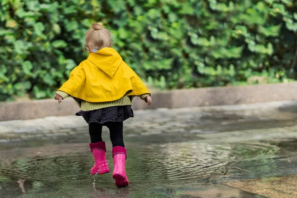 A nice little girl in a yellow raincoat and pink rubber boots runs on puddles with splashes and rejoices. Park, nature, outdoors. Universal Children's Day.