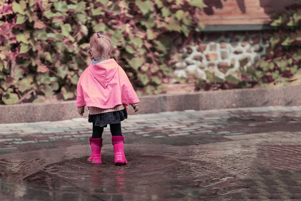 一个穿着玫瑰色雨衣和粉色橡胶靴的漂亮小女孩高兴地跳到水坑上 世界儿童节 免版税图库图片