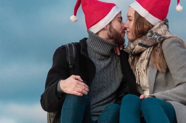 Paltolu ve kırmızı Noel şapkalı genç bir çift bulutlu ve dudaklarında öpücüklerle mavi bir gökyüzüne oturuyor. Romantik yeni yıl ilişkisi kavramı.