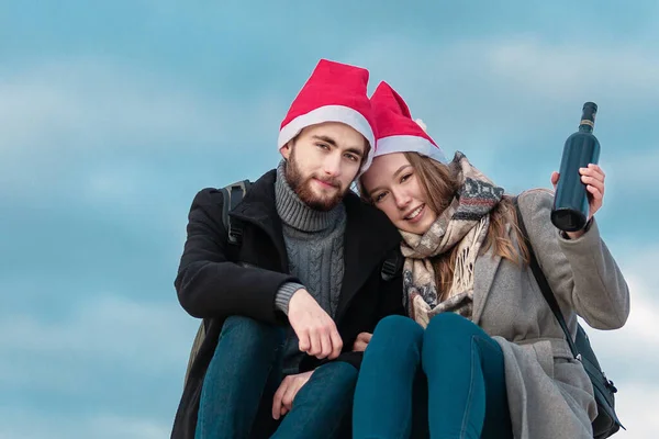 Paltolu ve kırmızı Noel şapkalı genç bir çift dışarıda oturuyor ve ellerinde bir şişe şarap tutuyorlar. Mutlu bir yeni yıl konsepti.